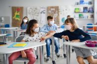 Kinder im Klassenzimmer, zwei Mädchen mit Maske berühren sich an den Ellbogen
